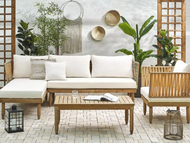 Odabir vrtnog namještaja - Moderan vanjski lounge set od svijetlog drva s bijelim jastucima, stolom, i dekoracijama okružen biljkama.
